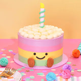 Jellycat Rainbow Birthday Cake with pastel layers, smiling face, and a candle on top by Crumbs & Doilies.